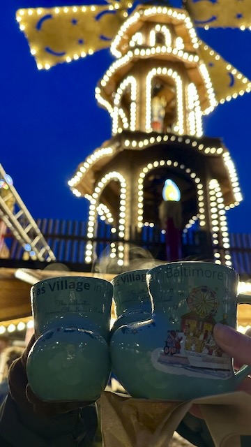 Two turquoise mugs with festive designs are held up in the foreground, featuring the text 'Christmas Village in Baltimore'. In the background, a brightly lit carousel structure is adorned with twinkling lights against a twilight sky.